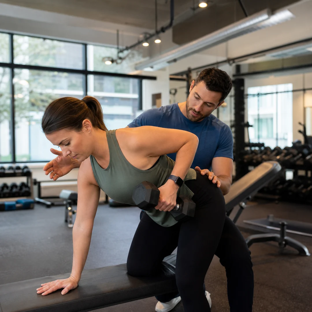 Modern spor salonunda antrenörün yetişkin üyeye dambılla kontrollü hareket yaptırdığı kare Instagram gönderisi